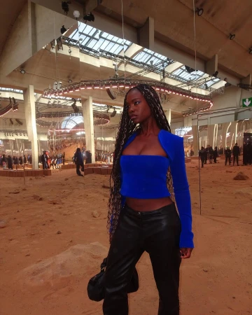 Fashionable Black woman in a bright blue top and black pants poses in a spacious, high-ceiling fashion show venue, exuding style.