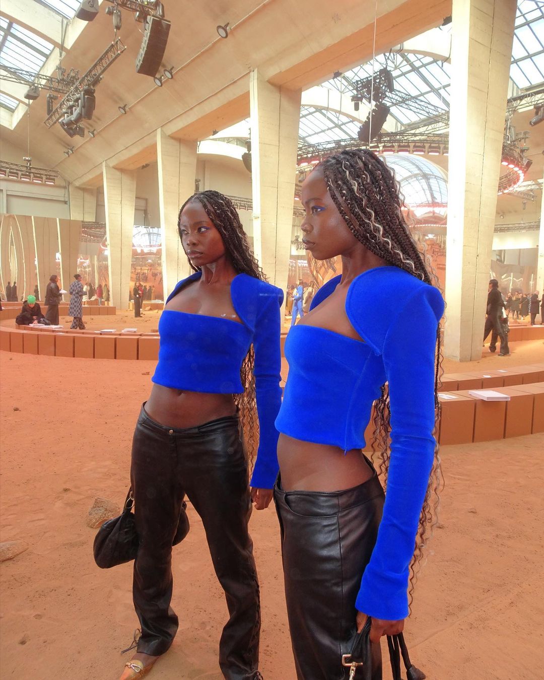 Two black women pose indoors wearing matching blue tops and black pants. Fashion runway setting visible. Long braids are apparent.