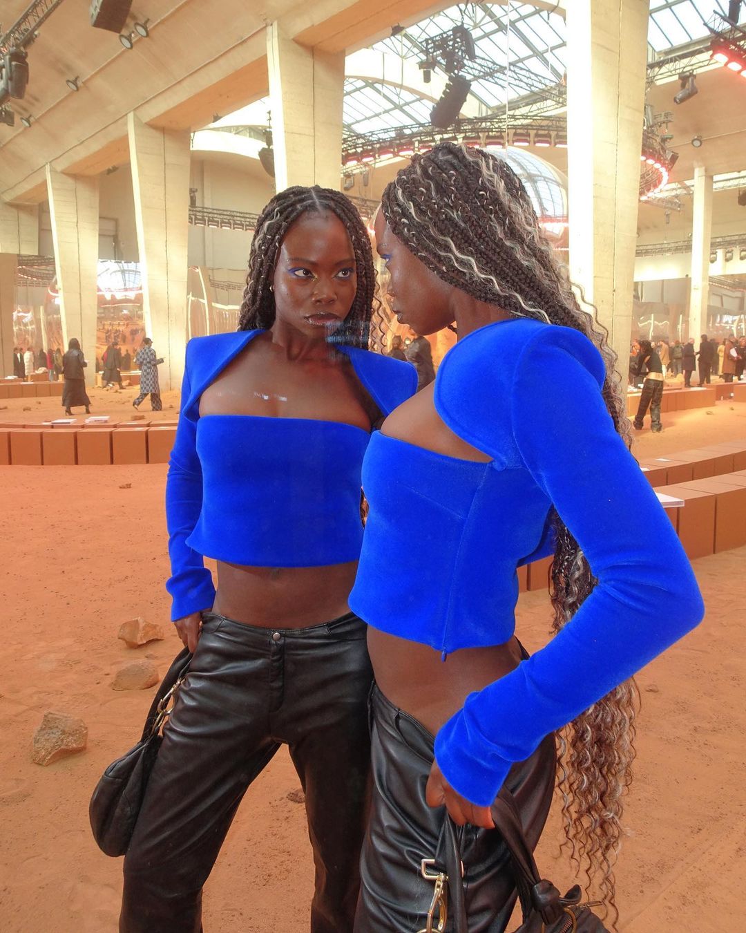 Two women in matching blue crop tops and black pants pose indoors, with one facing forward and the other in profile, for a fashion shot.