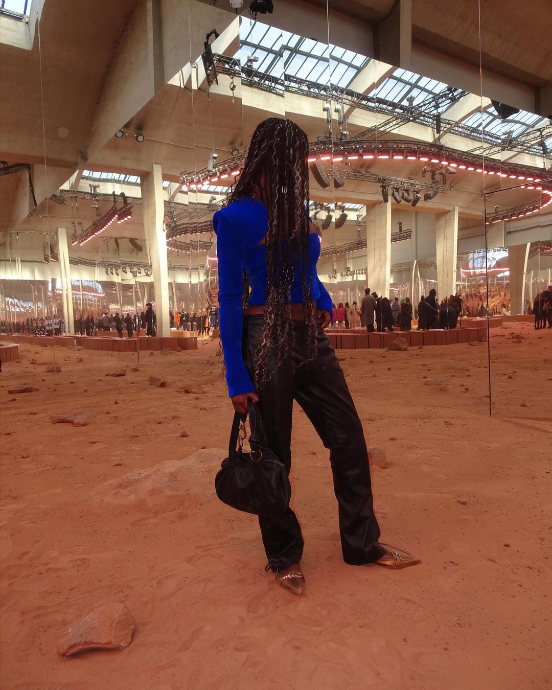 Runway fashion show featuring a Black model wearing a blue top, dark pants, heels, and carrying a black purse in a sandy venue.
