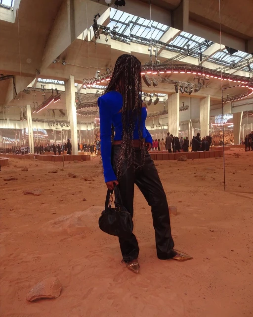 Runway fashion show featuring a Black model wearing a blue top, dark pants, heels, and carrying a black purse in a sandy venue.