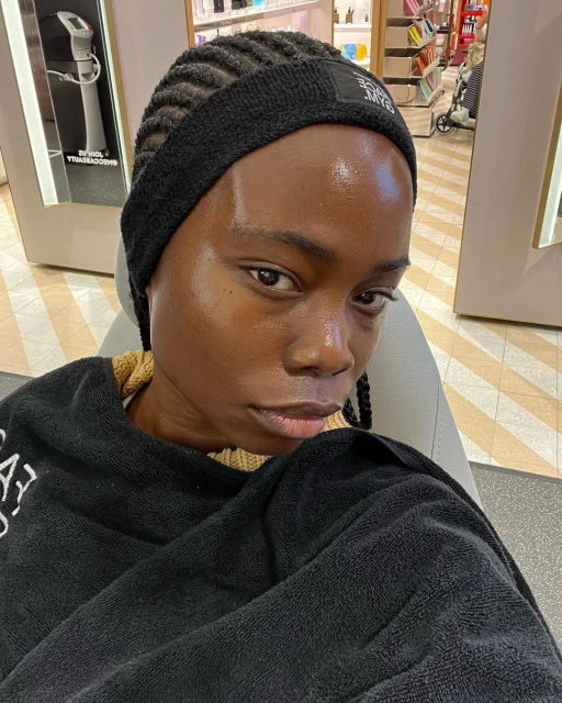Close-up portrait of a young Black woman with braided hair and a black headband, draped in a black towel, in a commercial environment.