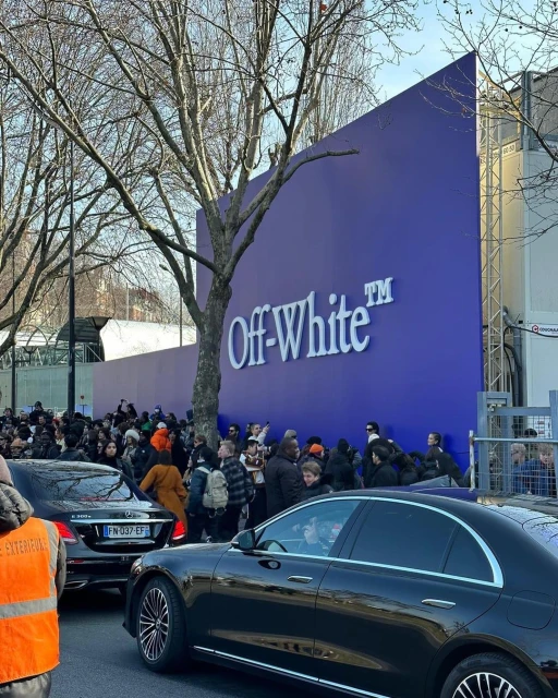 People gather near an Off-White store, with black cars parked in front and bare trees visible against a clear sky.
