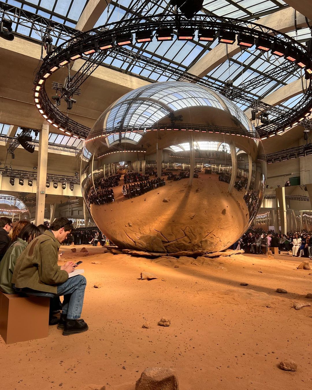 Fashion show setting: An indoor space with people seated around a giant silver sphere reflecting the scene, with catwalk and lights.
