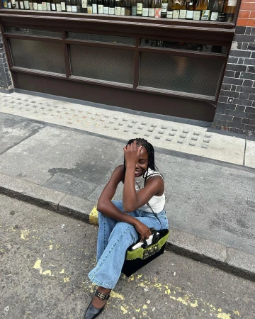 A Black woman seated on a stone curb wearing blue jeans and a white top, holding a black handbag with green accents.