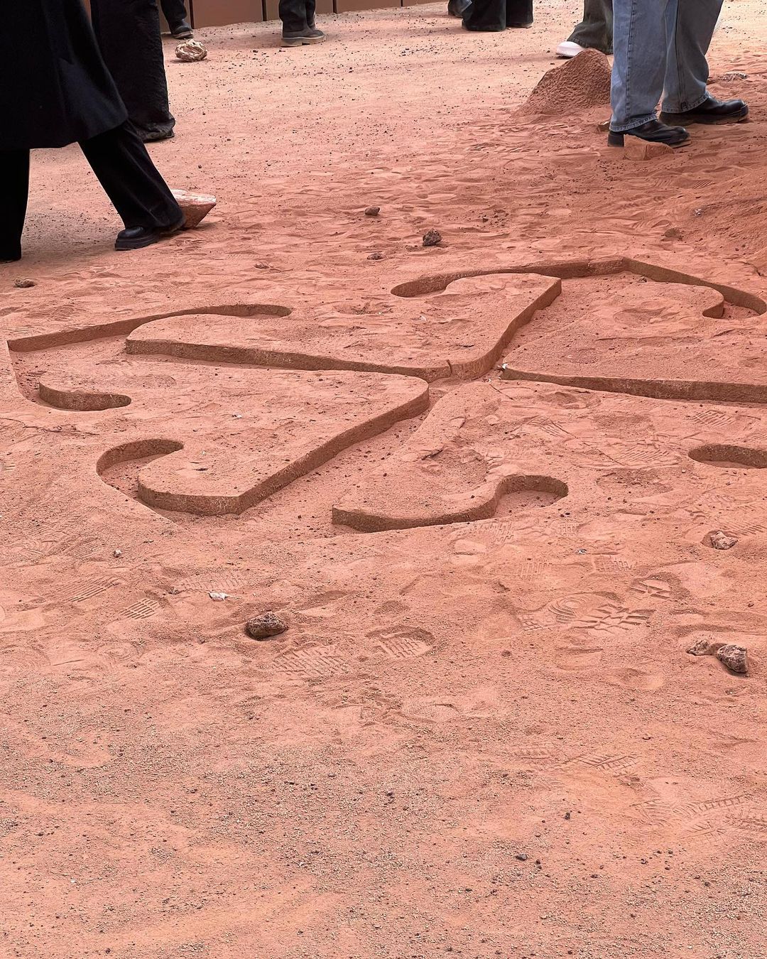Ground-level shot of a complex symbol carved into reddish-brown sand, surrounded by blurred figures of people standing around it.