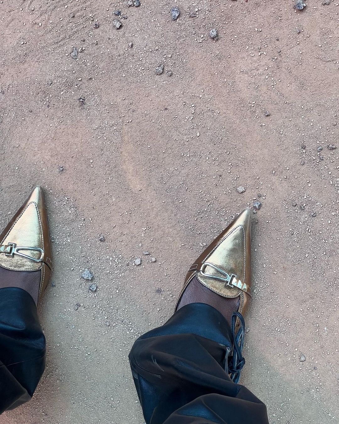 Feet wearing shiny gold pointed-toe heeled shoes and black leather pants stand on a ground of small rocks and sand.