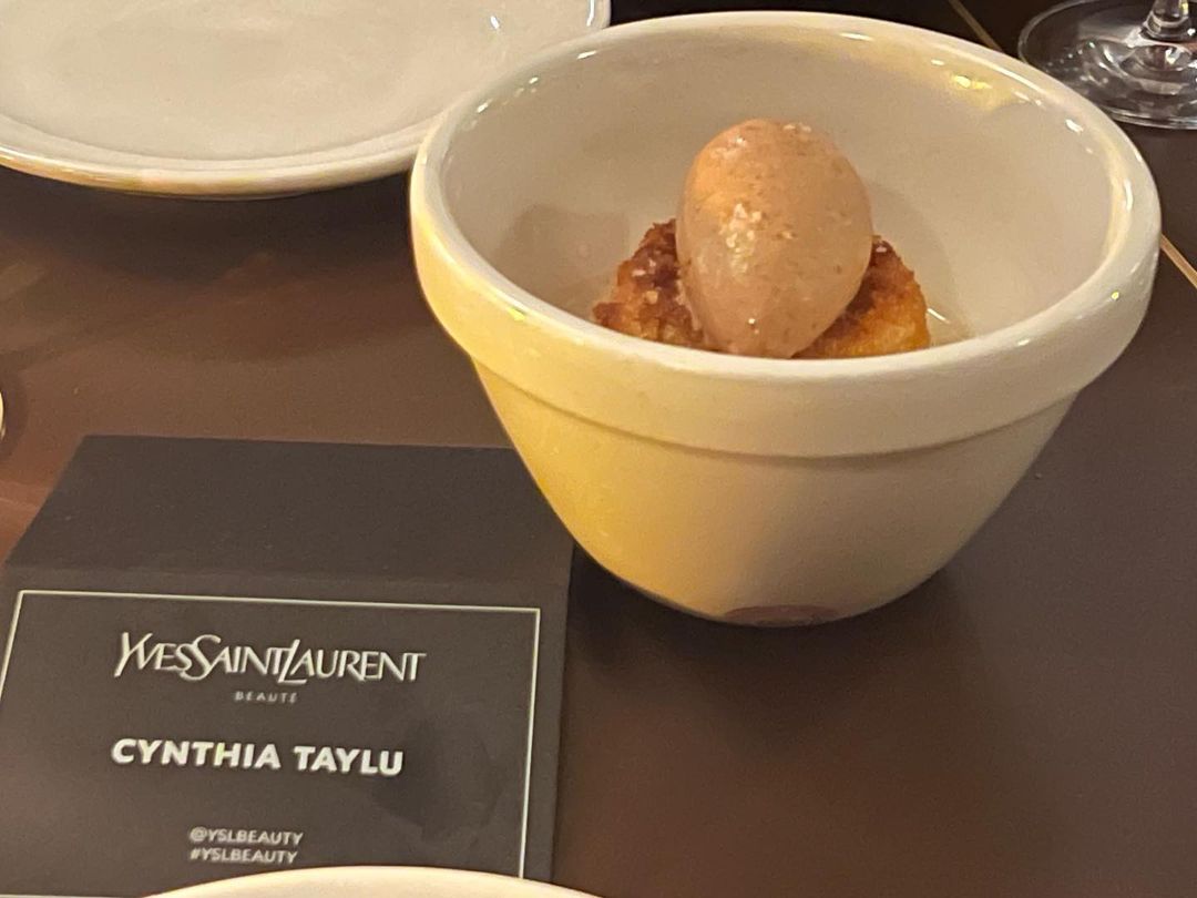 Dessert in a white bowl with a business card, featuring a scoop of ice cream or mousse on top of pastry on a brown table.