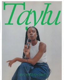 Black woman with braided hair wearing a white tank top and blue jeans holding a microphone against a plain white wall
