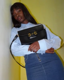 Black woman wearing a light blue collared shirt and a denim skirt holding a black leather handbag against a yellow wall.