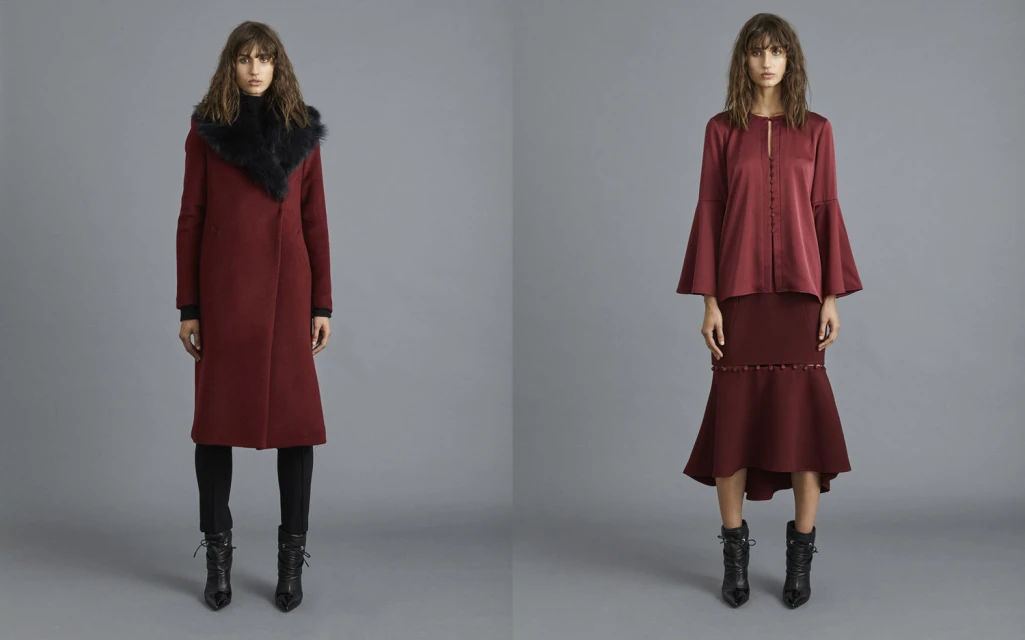 Two adult females wear fashionable burgundy overcoats and skirts in a studio with a gray background, complemented by black ankle boots.