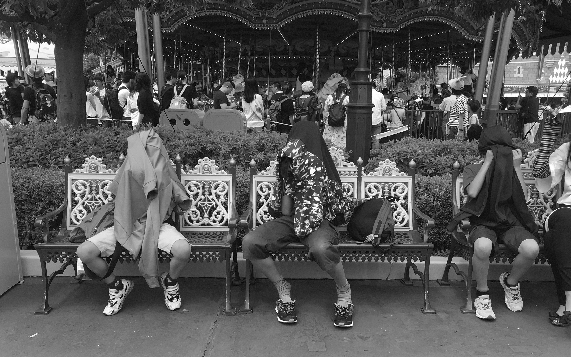 Monochrome shot captures three people resting on park benches in front of a carousel, their faces covered by shirts and jackets.