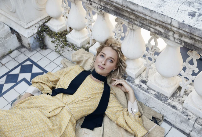 Blonde woman in a yellow dress and black scarf lies on a blanket on a blue-and-white tiled floor next to a white stone railing outdoors.