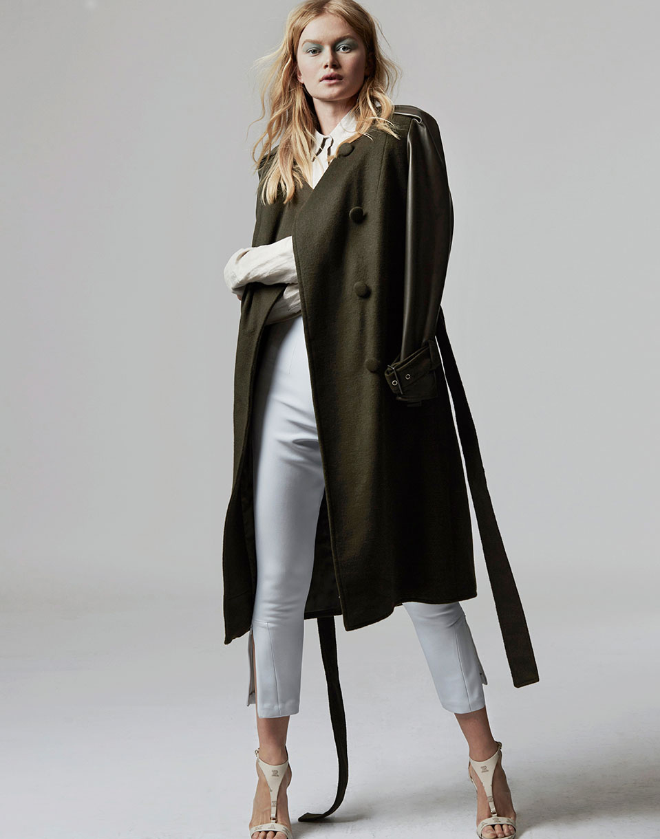 A young, blonde-haired woman poses in a green double-breasted trench coat, white top, and cropped pants against a neutral background.