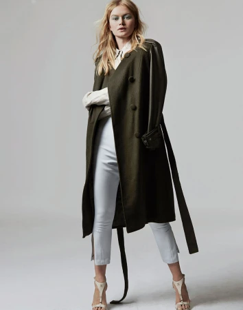 Model in olive green wool coat and white trousers standing against a seamless light background