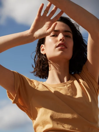 Woman in mustard yellow t-shirt with hands raised framing face against clear blue sky in Sydney, make-up by Andrea Black.