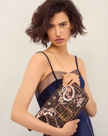 Woman with short dark wavy hair wearing a blue dress holding a patterned handbag against a plain background