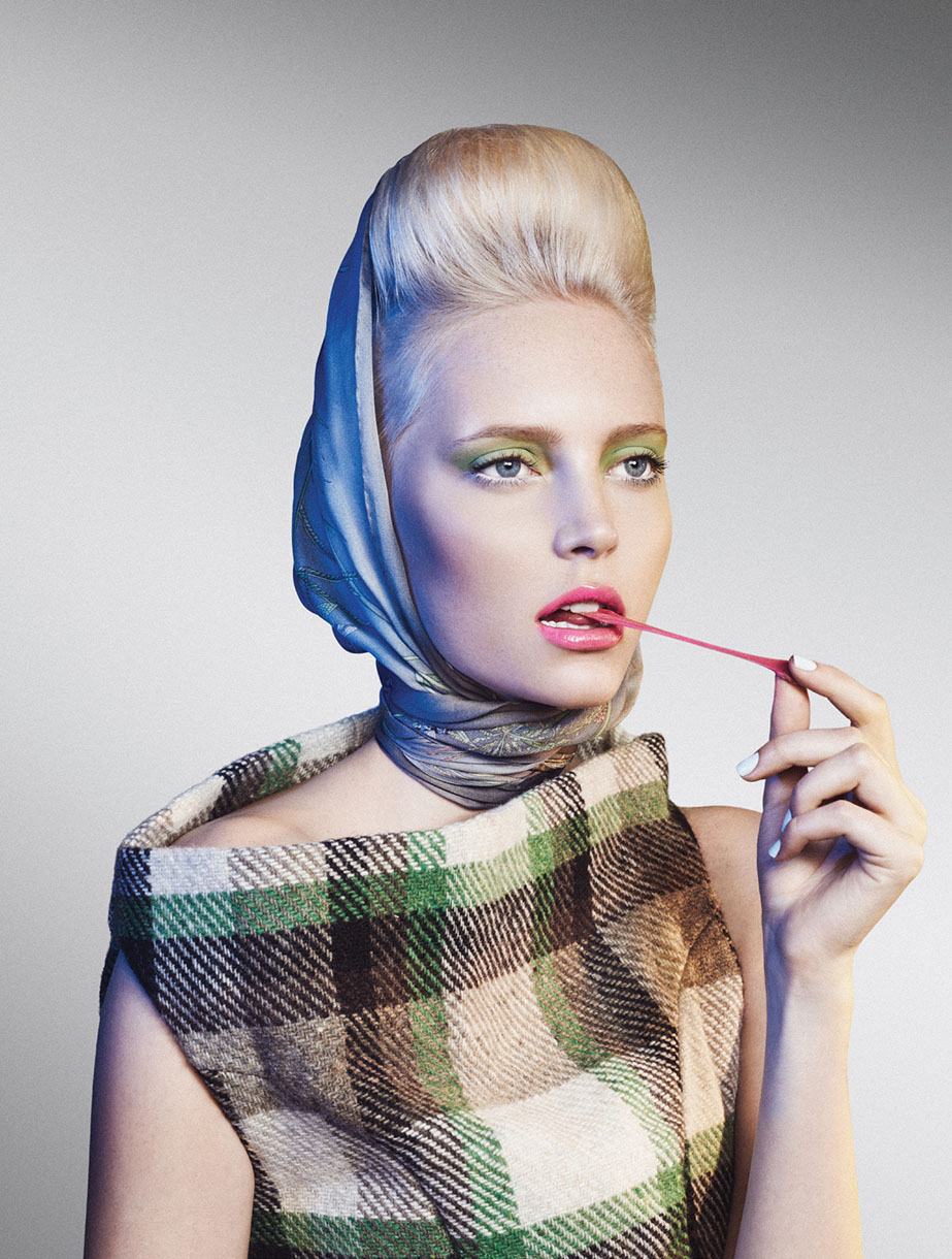 Woman in green plaid coat chews pink bubblegum with retro blonde updo hairstyle and headscarf in a studio fashion portrait.