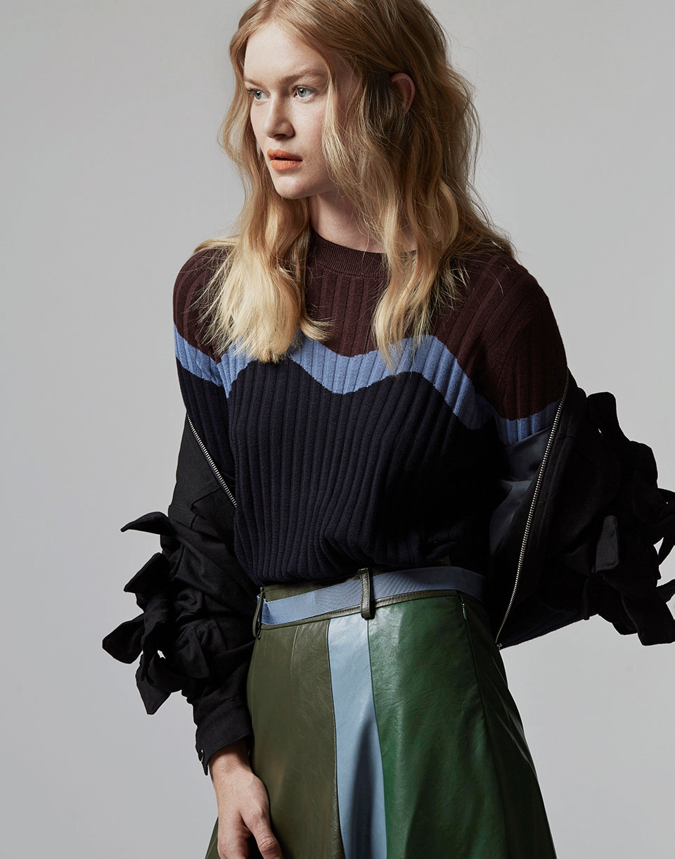 Blonde woman poses in an indoor studio. She wears a brown and navy blue sweater with a green, blue, and brown skirt with black jacket.