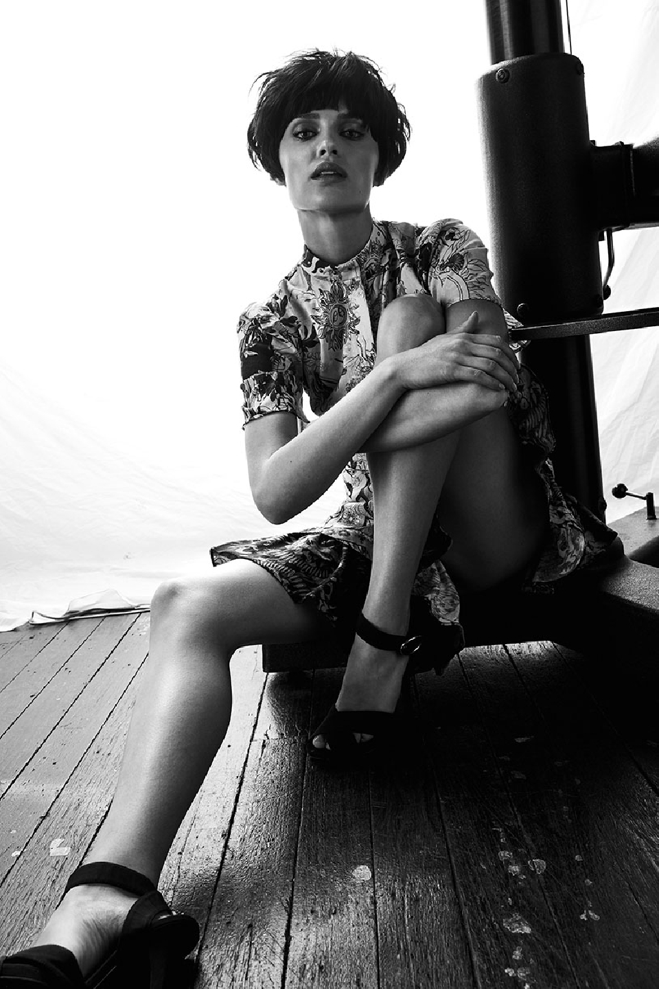 Monochrome portrait of a stylish woman in a patterned dress and black heels, seated on a wooden surface in front of a bright background.
