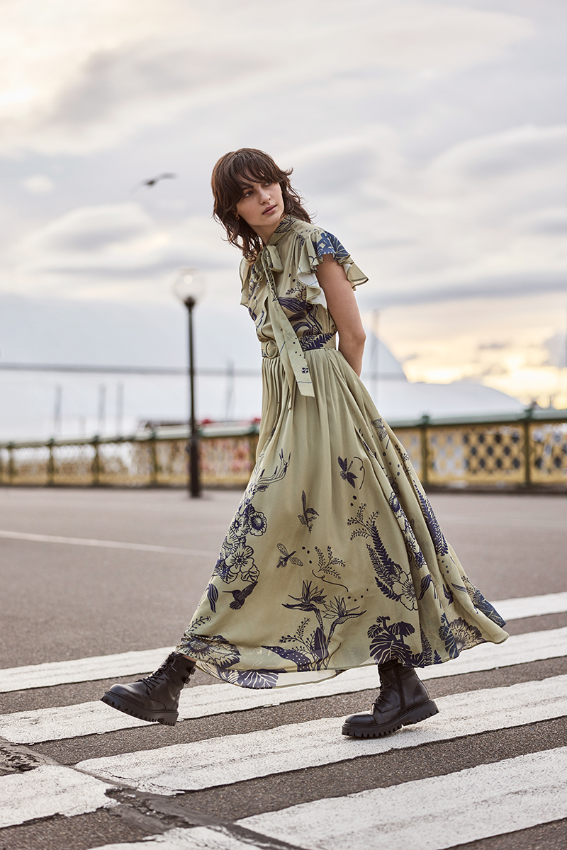 A woman walks on a crosswalk wearing a long, patterned dress and boots. The sky is cloudy and a building is visible in the background.