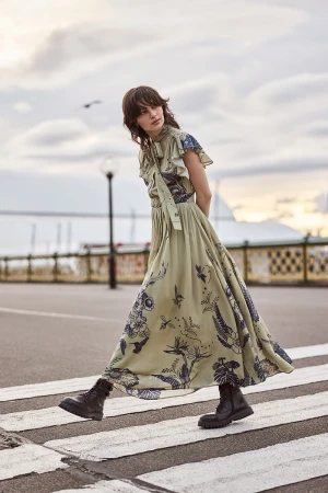 A woman walks on a crosswalk wearing a long, patterned dress and boots. The sky is cloudy and a building is visible in the background.