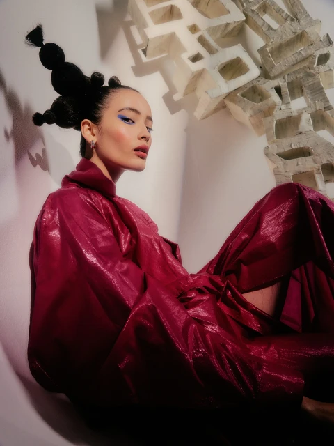Stylish woman in a vibrant magenta gown with unique black hair and makeup sits against a white background with abstract stone elements.