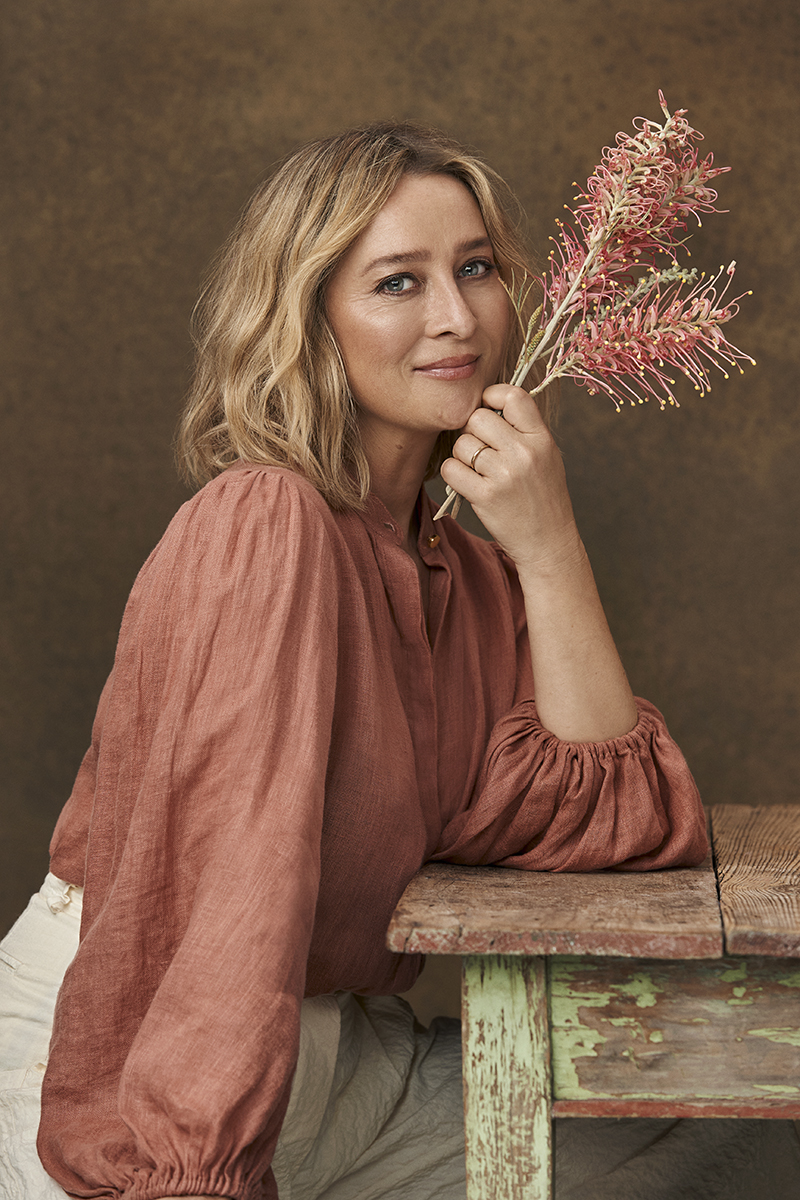 Portrait of adult woman with blond wavy hair, wearing a red top and holding pink protea flowers, leaning on a rustic wooden table.