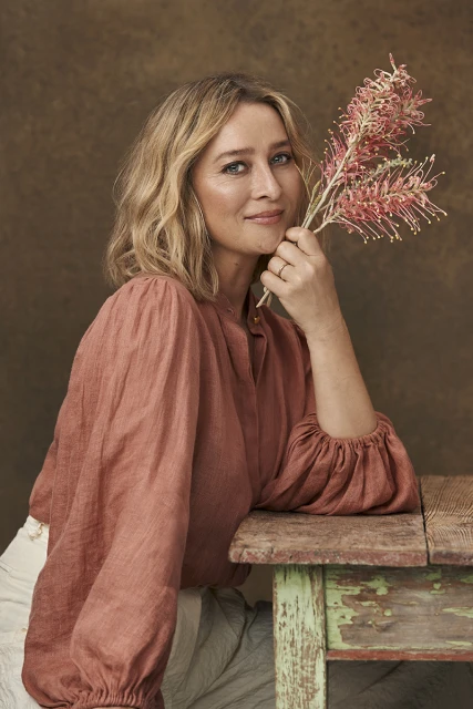 Portrait of adult woman with blond wavy hair, wearing a red top and holding pink protea flowers, leaning on a rustic wooden table.