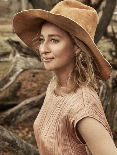 Portrait of a woman with blonde hair and a tan hat wearing a pleated peach-colored top, set against a blurry outdoor background.