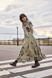 Model in patterned olive green midi dress walking on a crosswalk with dark combat boots under a cloudy sky.
