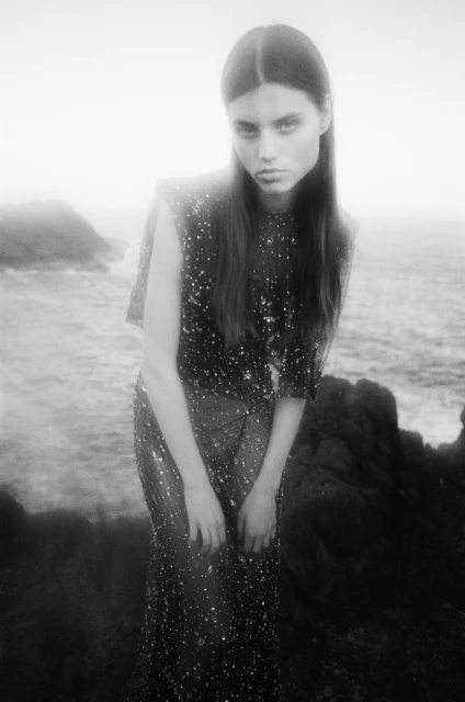 Black and white portrait of a young woman in a sequined dress standing on rocky ground near the ocean under a foggy sky.