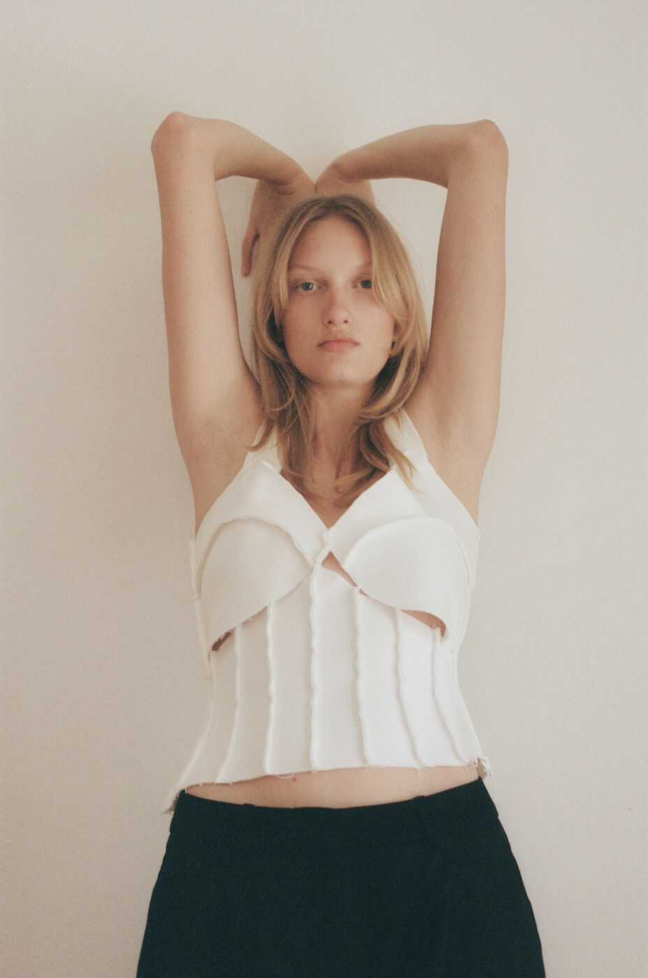 Young woman with blonde hair and a white top poses against a neutral background with her arms above her head in a studio shot.