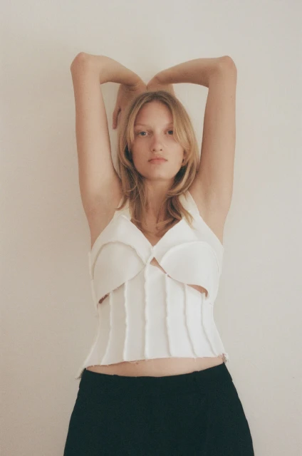 Young woman with blonde hair and a white top poses against a neutral background with her arms above her head in a studio shot.