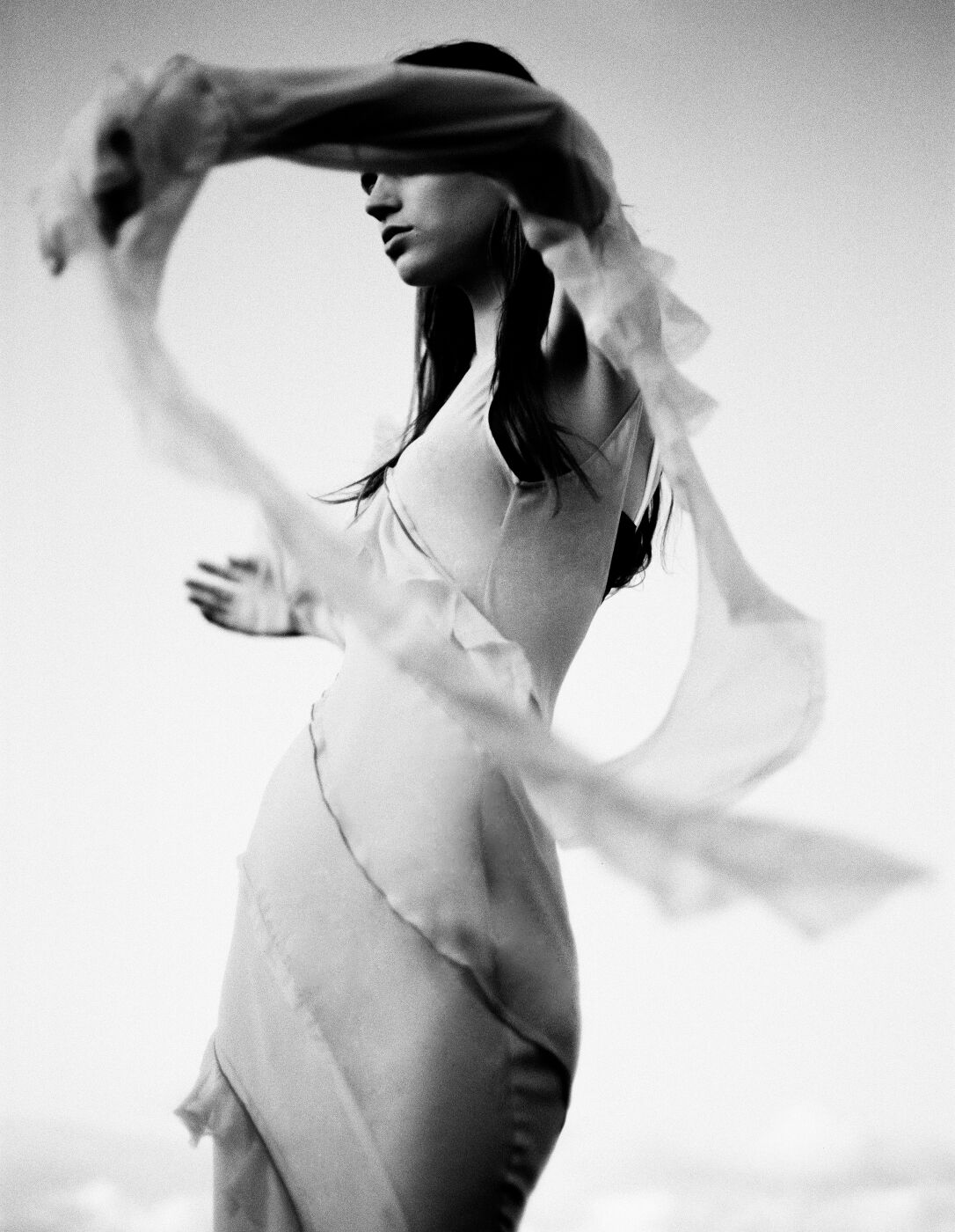 Monochrome portrait of a young woman in flowing dress, face partially obscured by translucent fabric, evoking ethereal fashion photography.