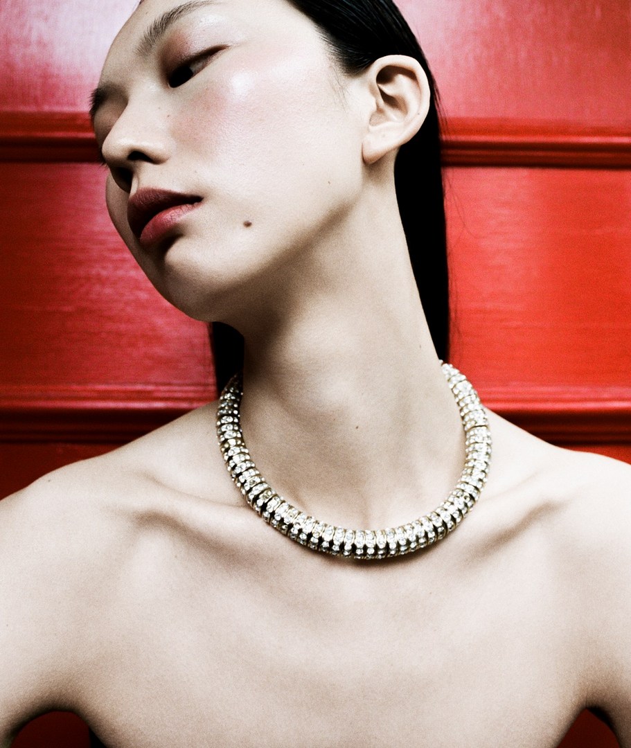 Close-up studio shot of a young Asian woman with porcelain skin wearing a statement silver choker necklace against a red wall.