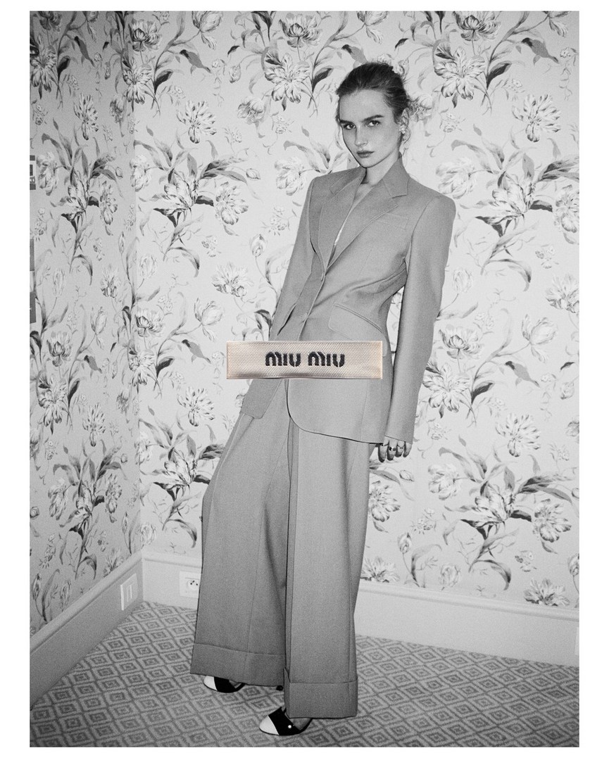 Monochrome shot of a woman in a suit posing in front of a floral patterned wall, showcasing fashion and indoor studio-like aesthetics.