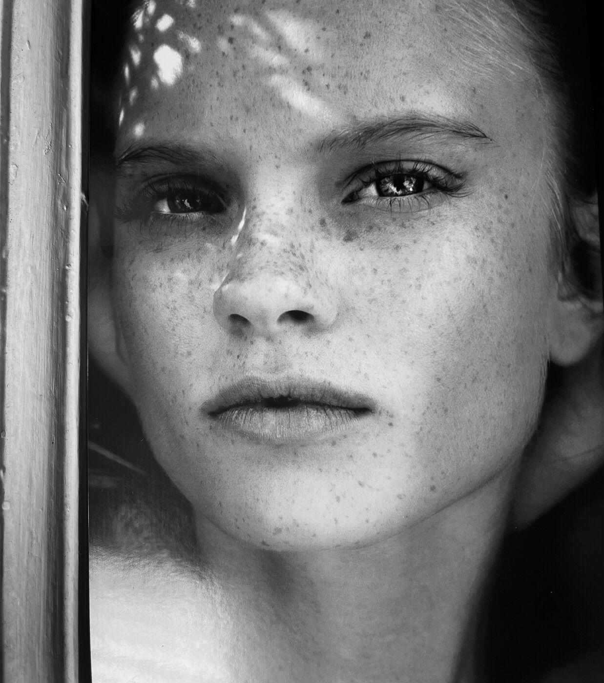 Monochrome portrait of a light-skinned woman with freckles, looking directly at the camera with neutral expression and soft, dappled lighting.