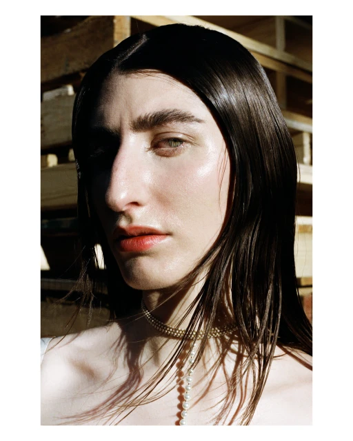 Portrait of a young adult woman with dark hair, fair skin, and orange lipstick, wearing a pearl necklace against a wooden background.