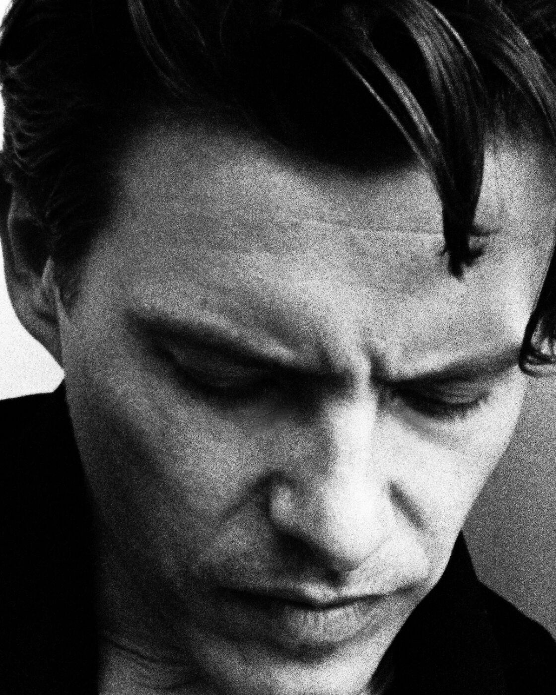 Monochrome portrait of an adult male with dark hair, gazing downwards in a close-up shot with a blurred, nondescript background.