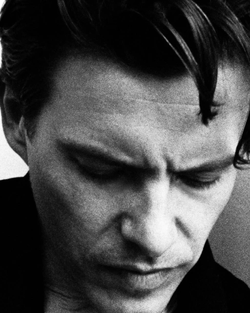 Monochrome portrait of an adult male with dark hair, gazing downwards in a close-up shot with a blurred, nondescript background.