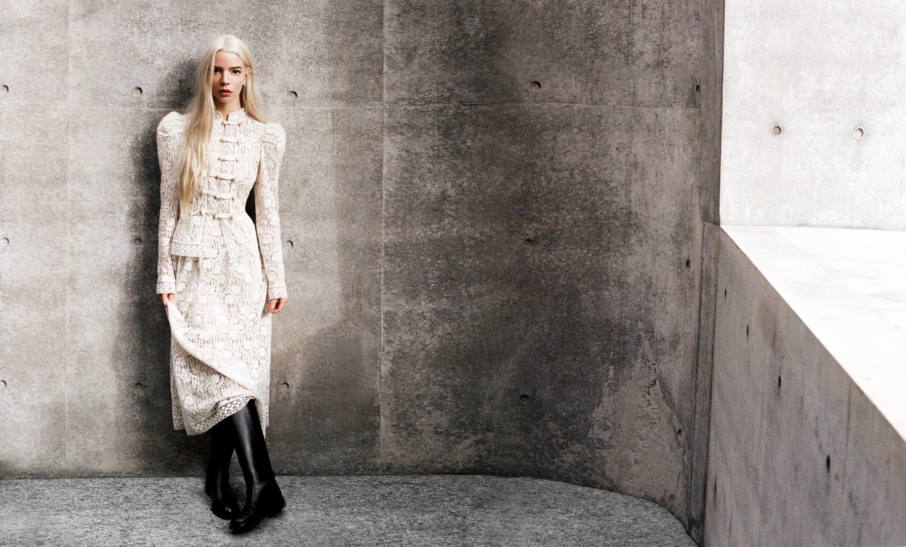 Anya Taylor-Joy poses in a cream-colored, lacy dress and black boots against a textured concrete wall in a fashion photography setting.