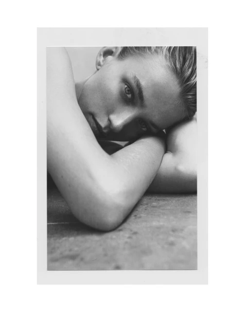 Monochrome portrait of a young fair-skinned person with light eyes, head tilted and resting on their arms, against a blurred background.