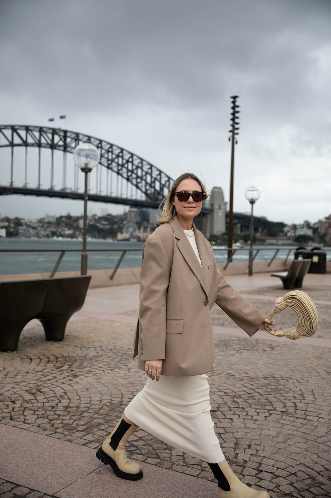 A woman walks on a waterfront promenade with the Sydney Harbour Bridge behind her, wearing a blazer, dress, boots and carrying a bag.