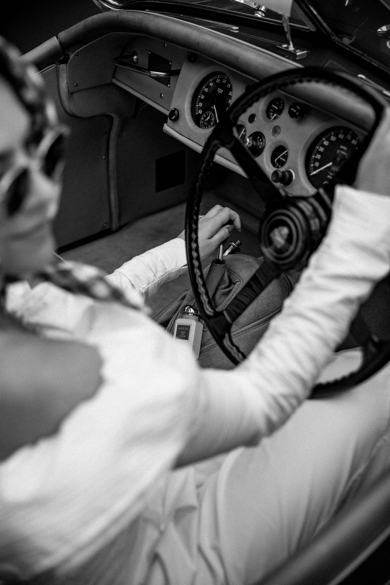A woman driving a vintage car, shown in black and white, wearing stylish sunglasses and gloves, with a classic dashboard visible.