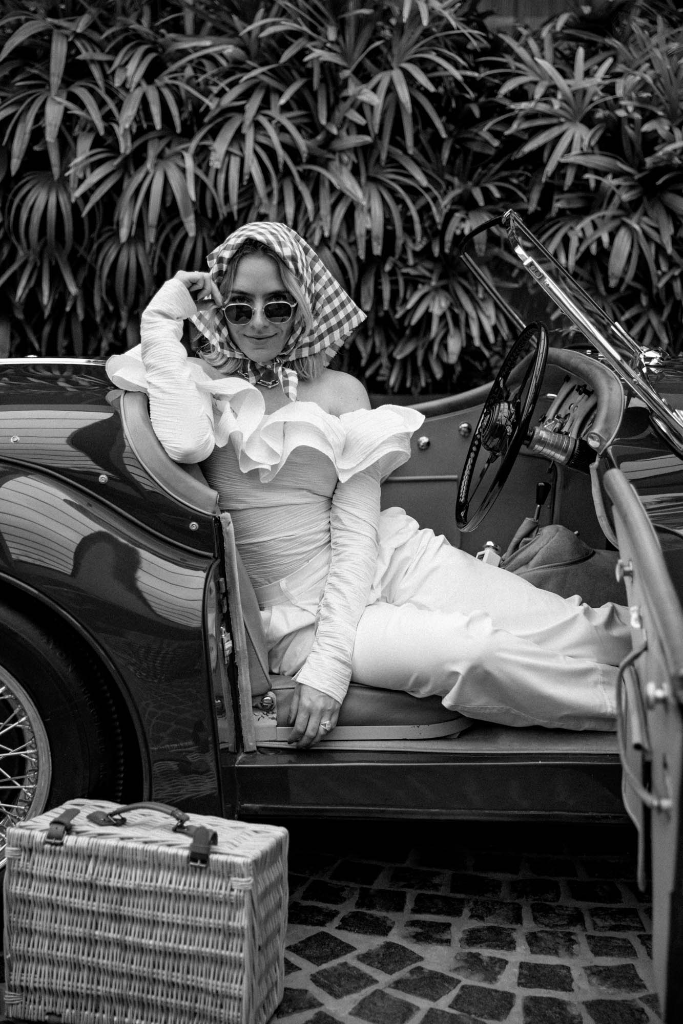 Elegant woman in a vintage convertible car wearing a headscarf, sunglasses, and a dress with ruffled sleeves, accompanied by a wicker picnic basket.