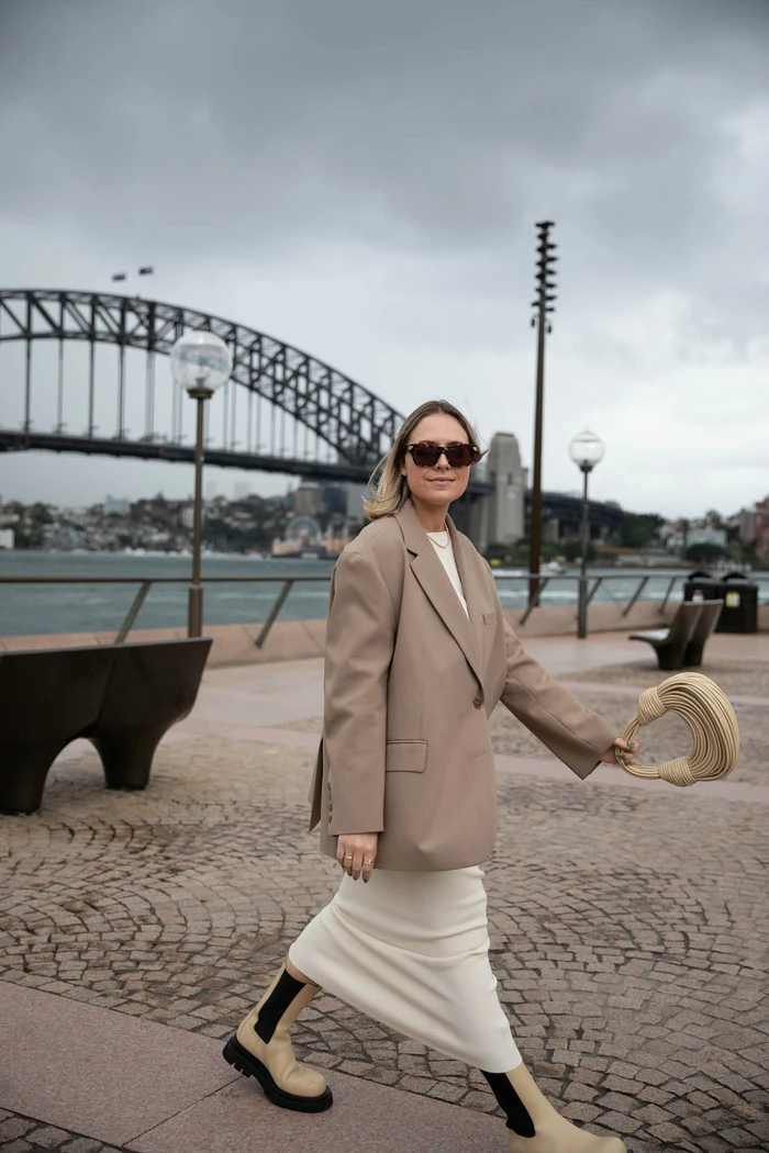 Woman walking on a paved waterfront in Sydney wearing a beige blazer, white skirt, and boots with the Sydney Harbour Bridge behind.