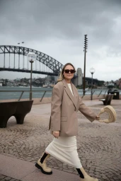 Woman walking on a paved waterfront in Sydney wearing a beige blazer, white skirt, and boots with the Sydney Harbour Bridge behind.