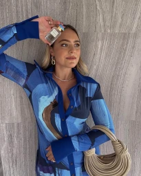 Woman in blue printed shirt holding a perfume bottle and beige handbag against a stone wall