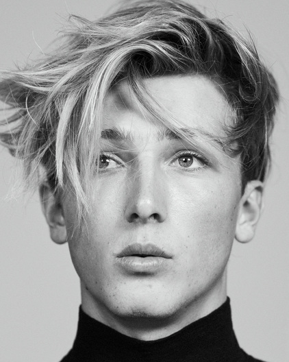 Headshot of a young adult with light hair and a black turtleneck against a gray background, showcasing a modern, minimalist portrait.
